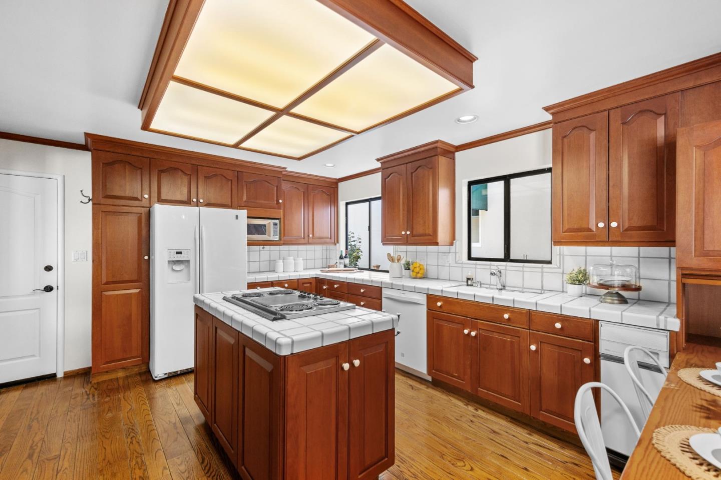 Kitchen with reddish-brown wood cabinets, tiled counters, central cooktop island, and hardwood floors.