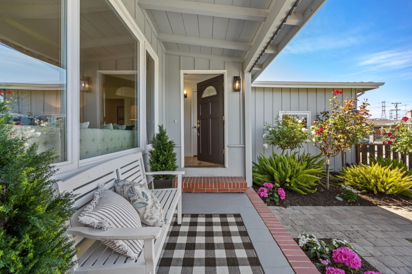 Front porch with a dark entry door, white cushioned bench, potted plants, and flowering roses.