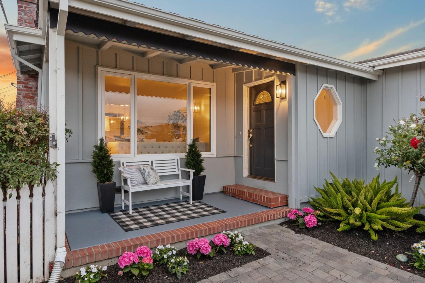 Gray home’s covered front porch with a dark entry door, white bench, brick steps, and colorful flowers.
