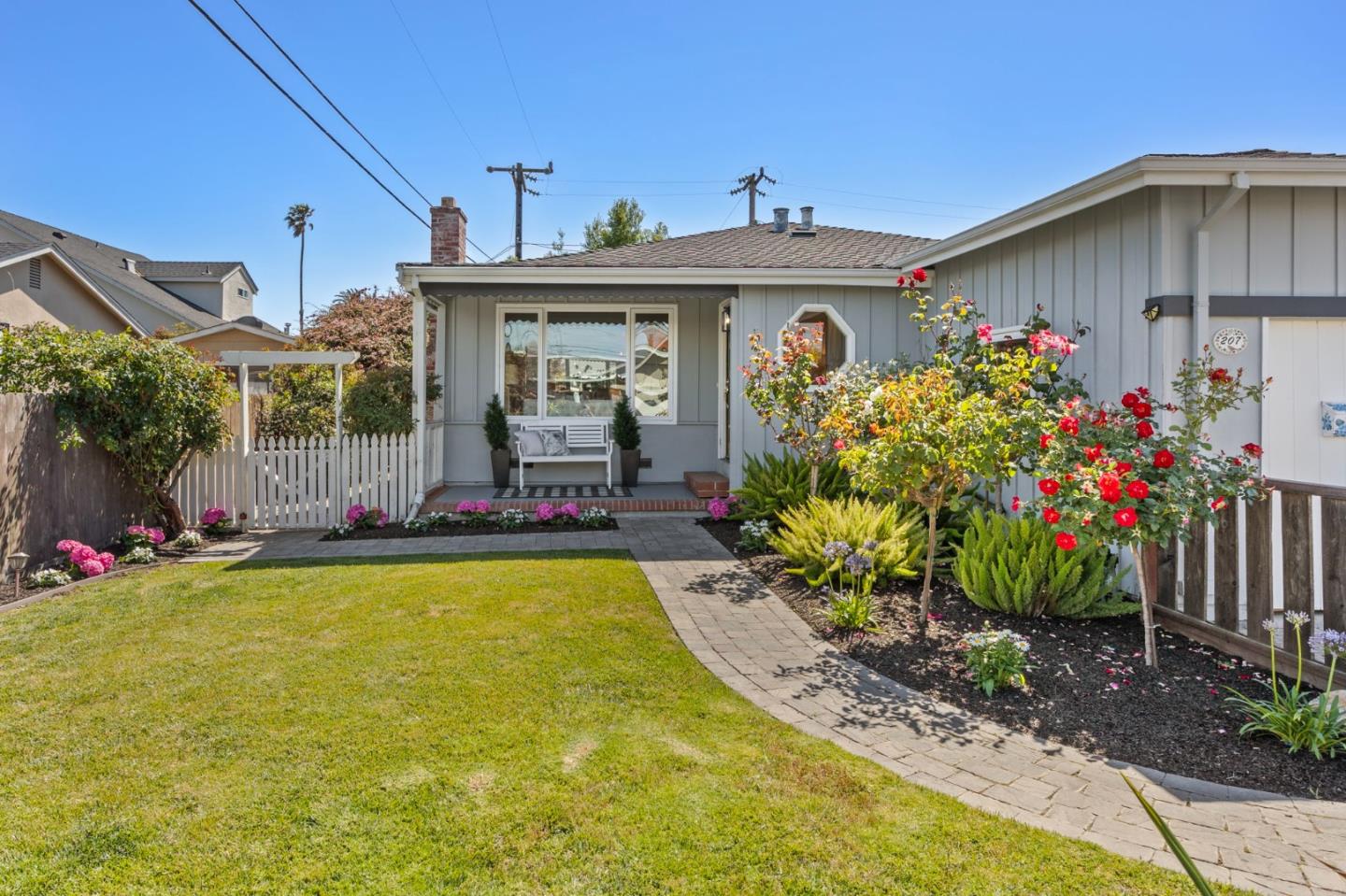 Gray home with a covered porch, curved garden walkway, red roses, and white picket gate.