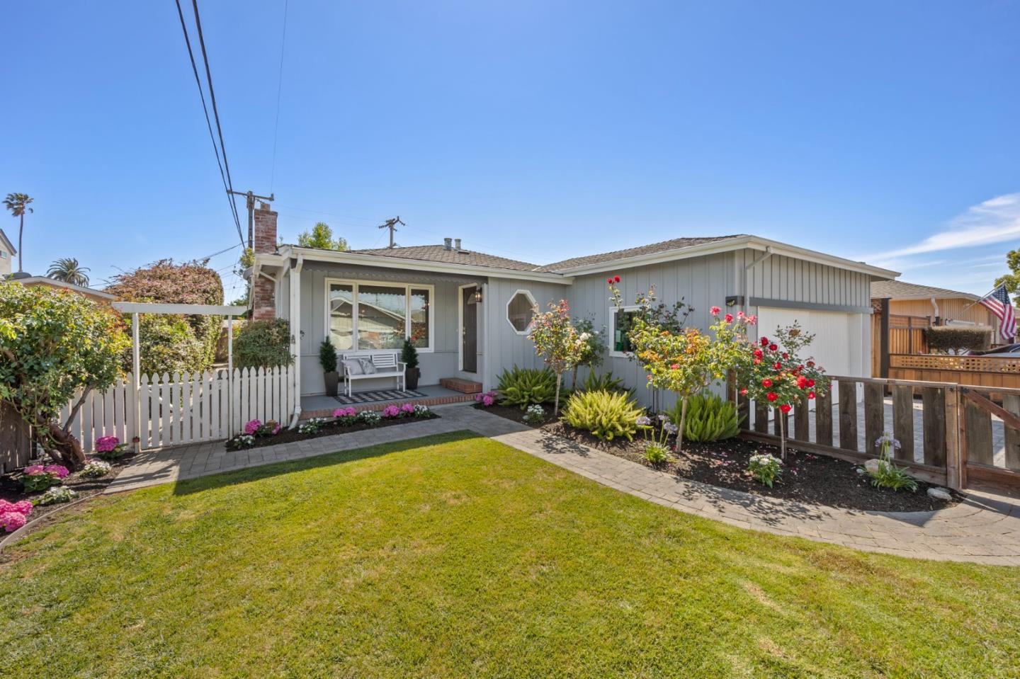 Single-story gray home with a white garage, flowering garden, picket fence, and broad front lawn.