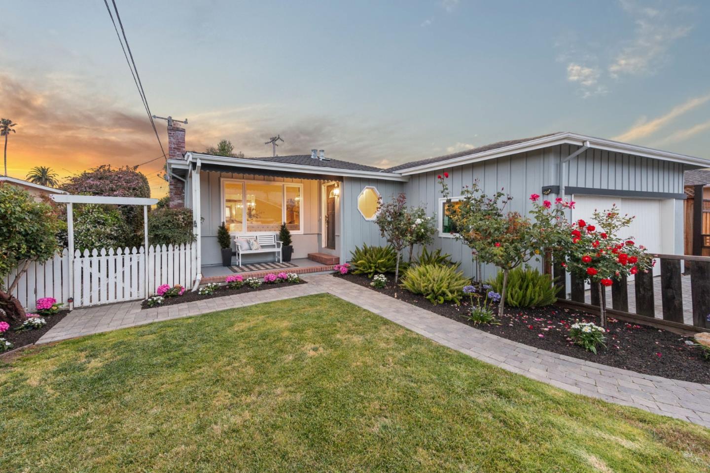 Gray single-story home with a white picket gate, flower beds, and a brick path at sunset.