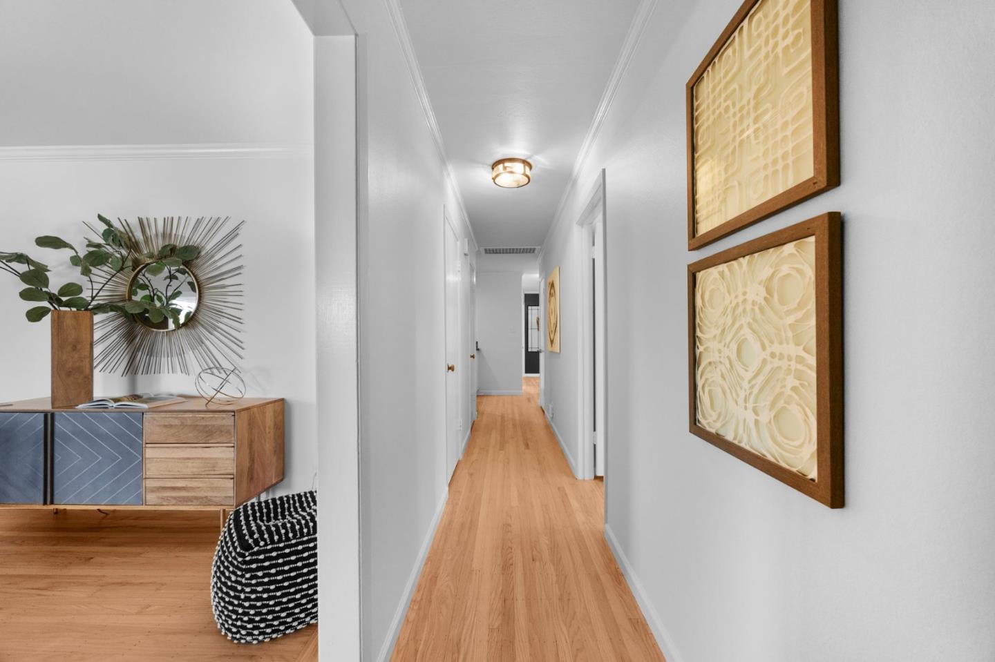 Bright hallway with hardwood floors, framed artwork, and a wooden console beside a sunburst mirror.