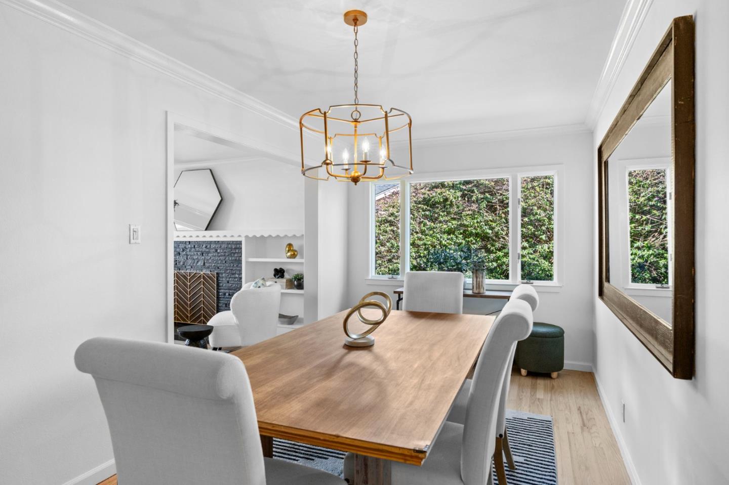 Dining room with a light-wood table, white chairs, brass chandelier, and windows overlooking greenery.