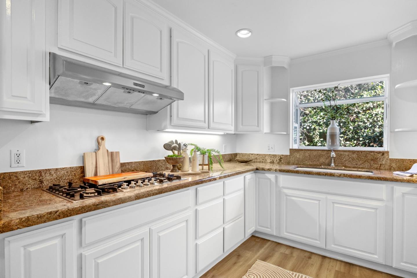 Bright kitchen with white paneled cabinets, brown stone counters, and a sink below a window overlooking greenery.