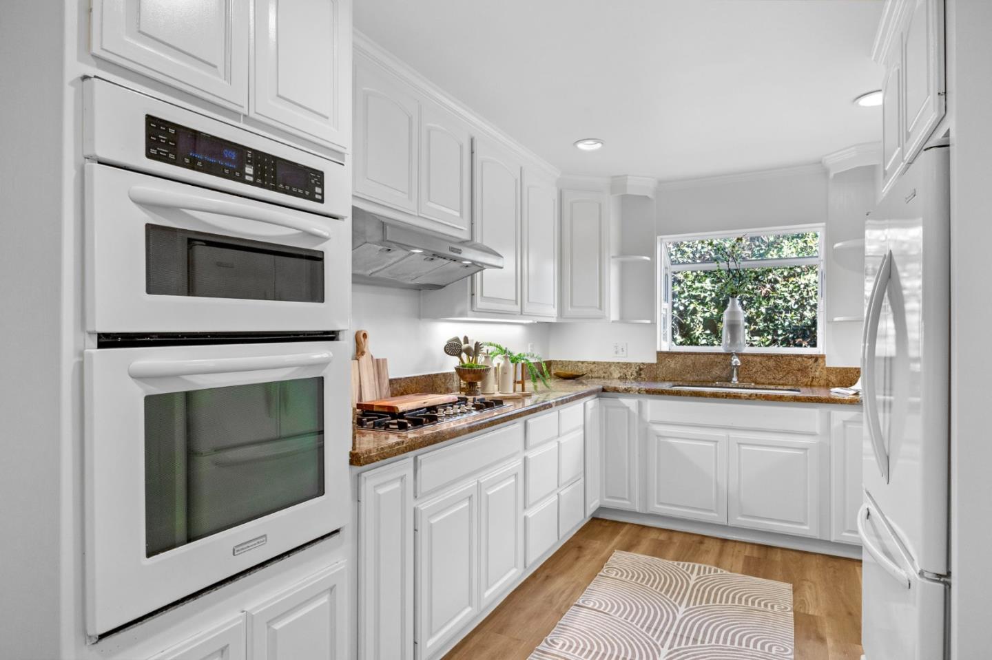 Bright kitchen with white cabinetry, double wall oven, and brown stone counters beside a window.