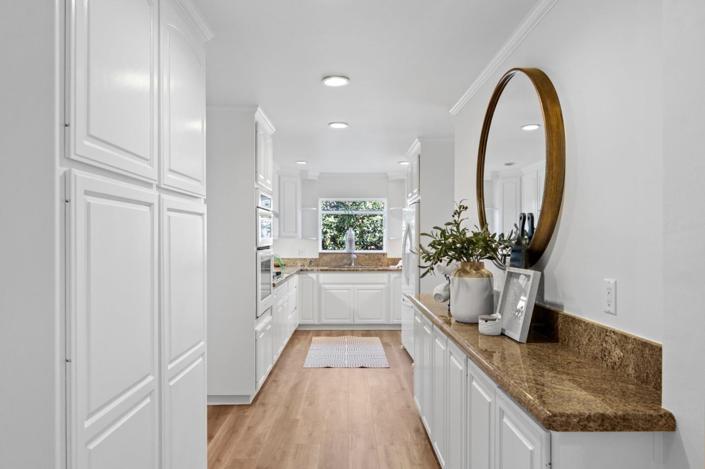 Bright galley kitchen with white cabinets, brown granite counters, and light wood floors.