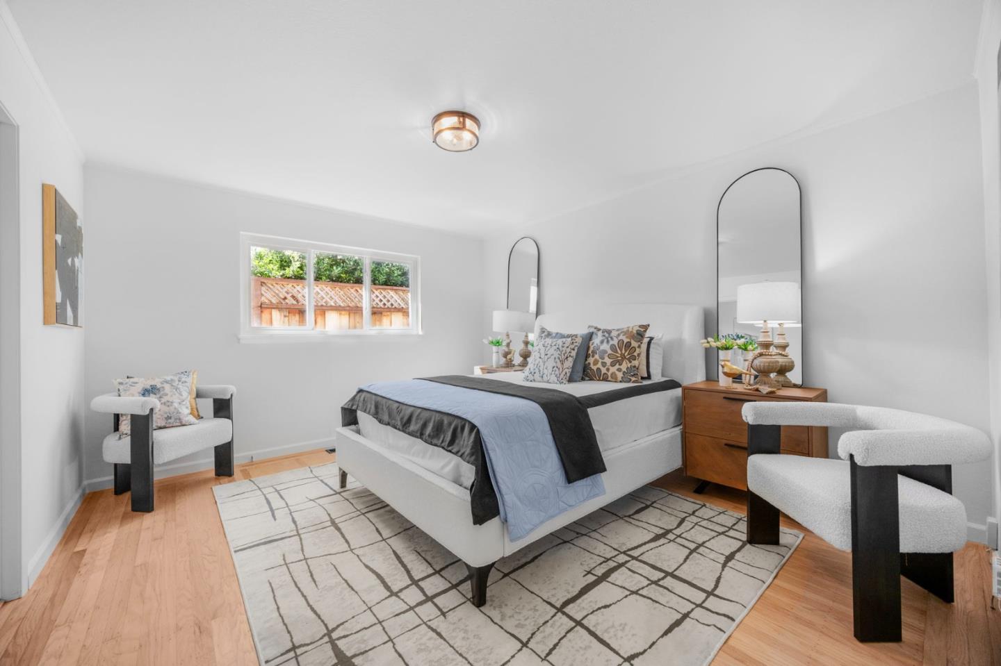 Staged bedroom with a white bed, arched mirrors, upholstered chairs, and a window overlooking a fenced garden.