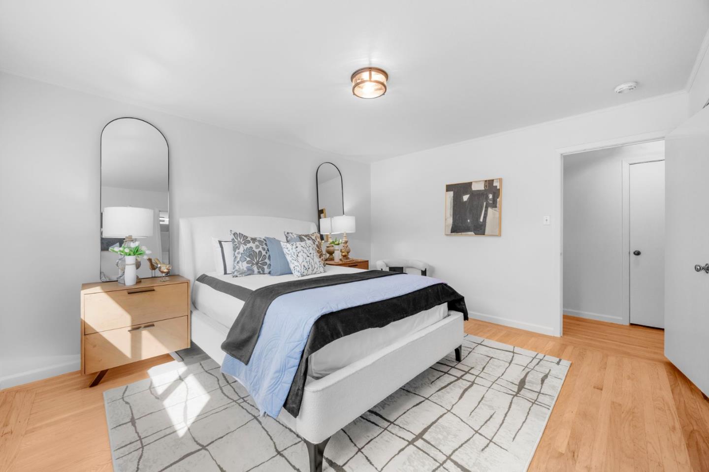 Staged bedroom with a white upholstered bed, blue and gray bedding, arched mirrors, and hardwood floors.