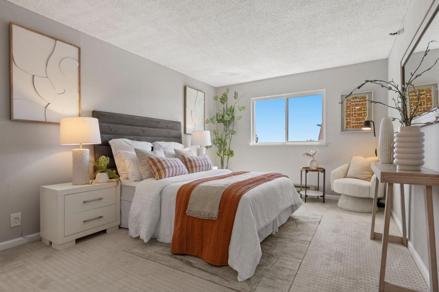 Staged bedroom with a gray headboard, white bedding, rust-colored throw, and window-side sitting area.