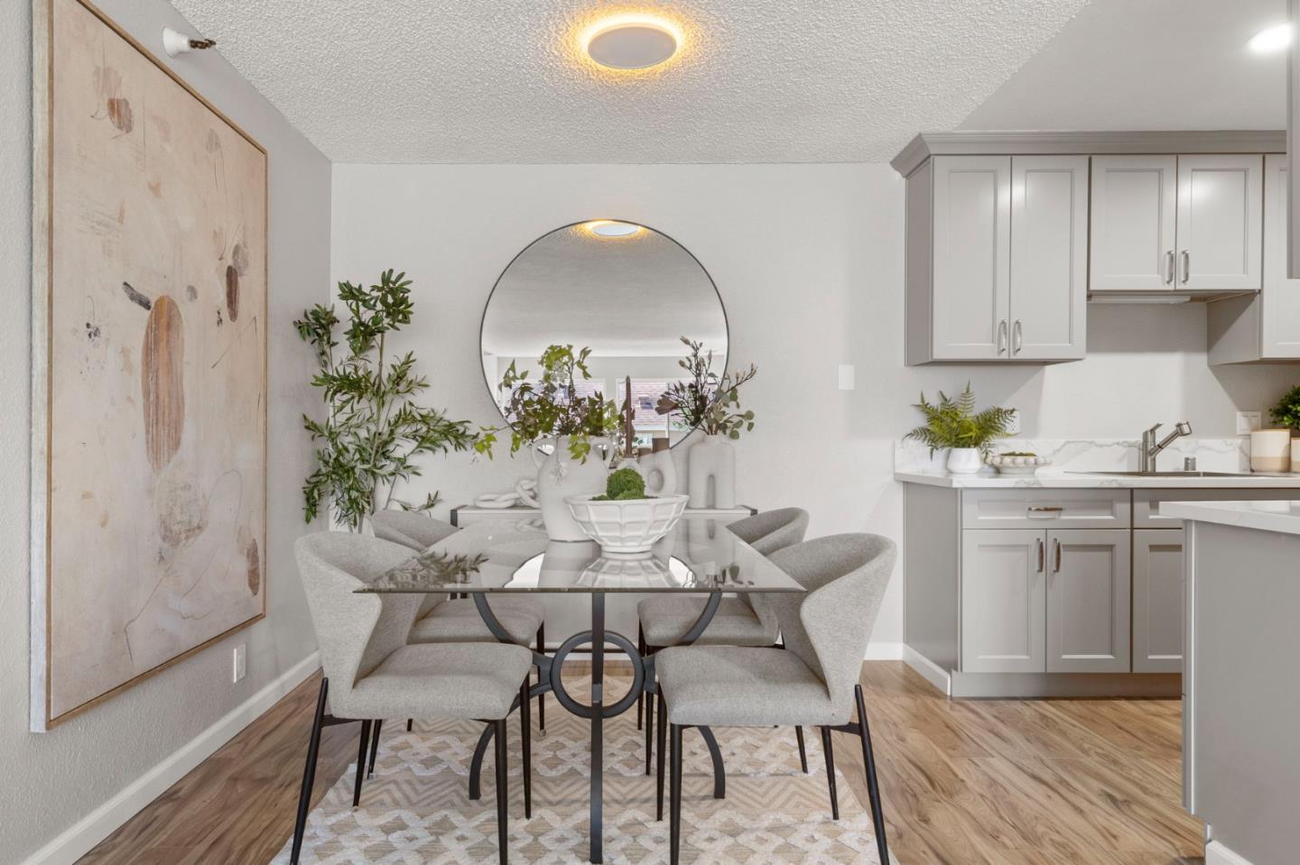 Dining area with a glass table, gray chairs, round mirror, and white kitchen cabinetry.