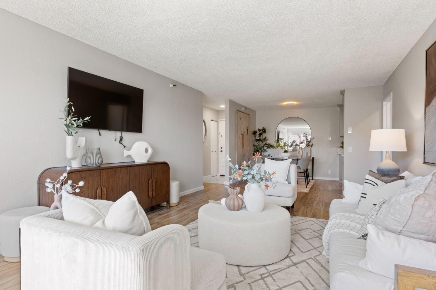 Bright living room with white sofas, a round ottoman, and a dining area beyond.