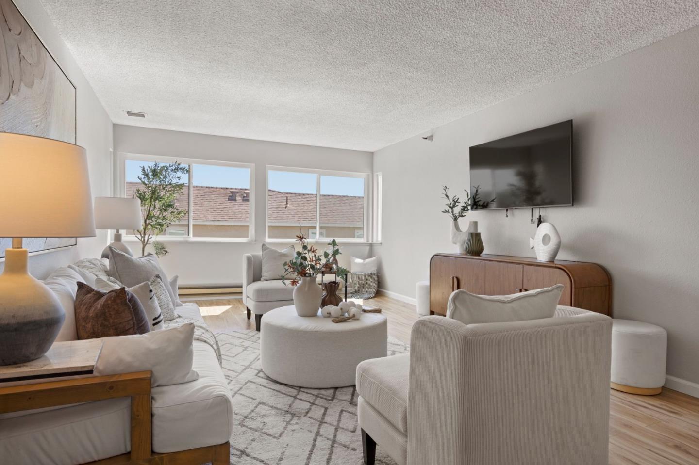 Staged living room with white seating, a round ottoman, and a wall-mounted television.