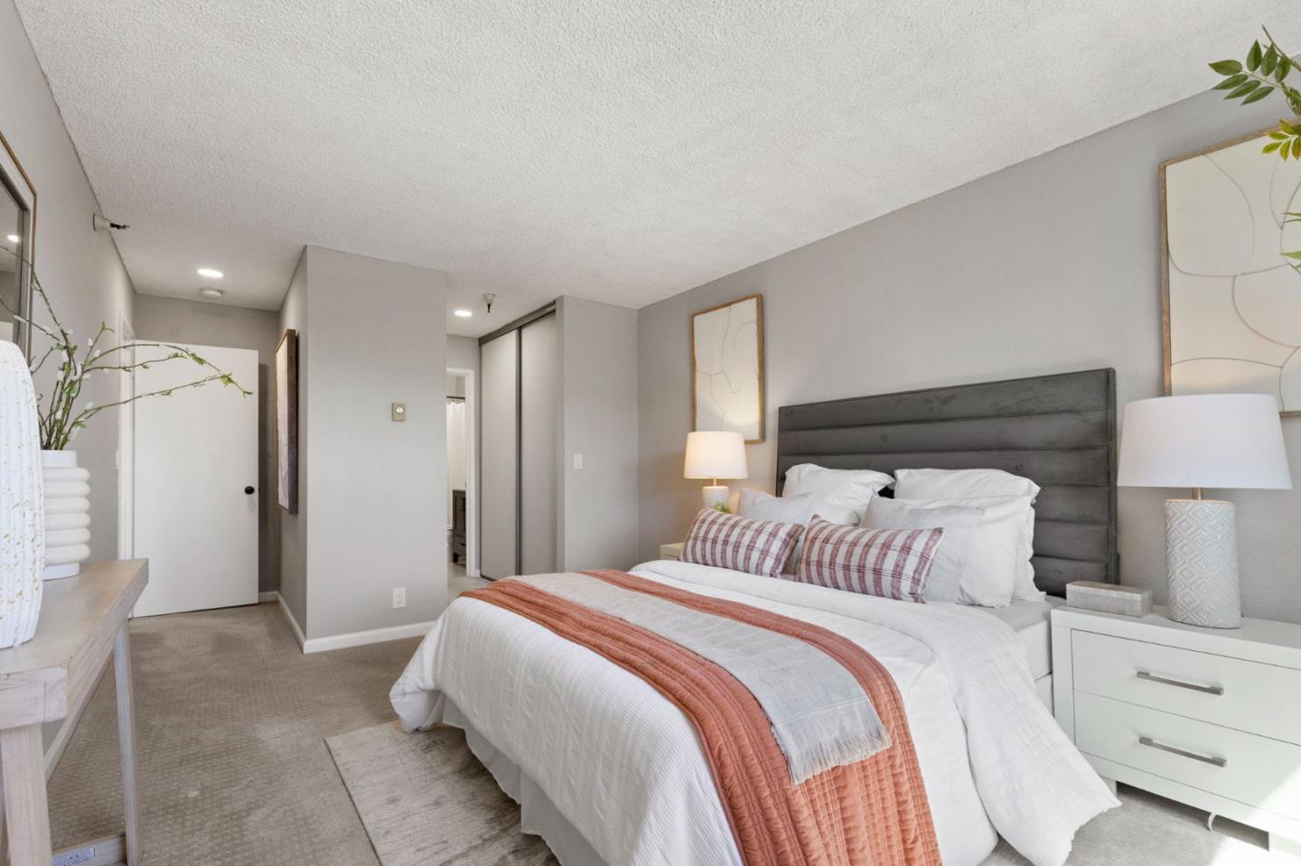 Staged bedroom with a large white bed, rust-colored throw, and dark upholstered headboard.