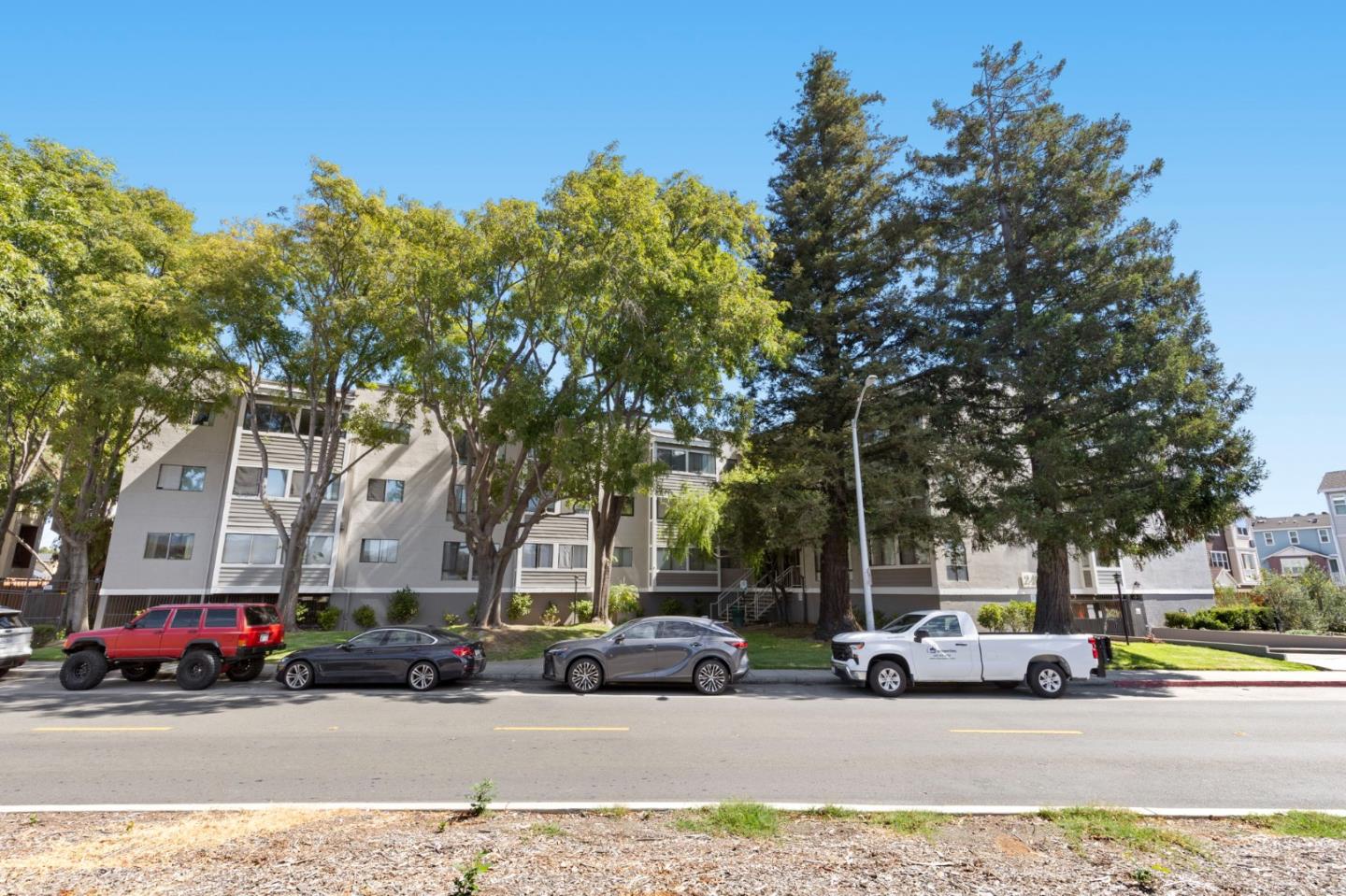 Condominium building behind mature trees, with parked cars along the street.
