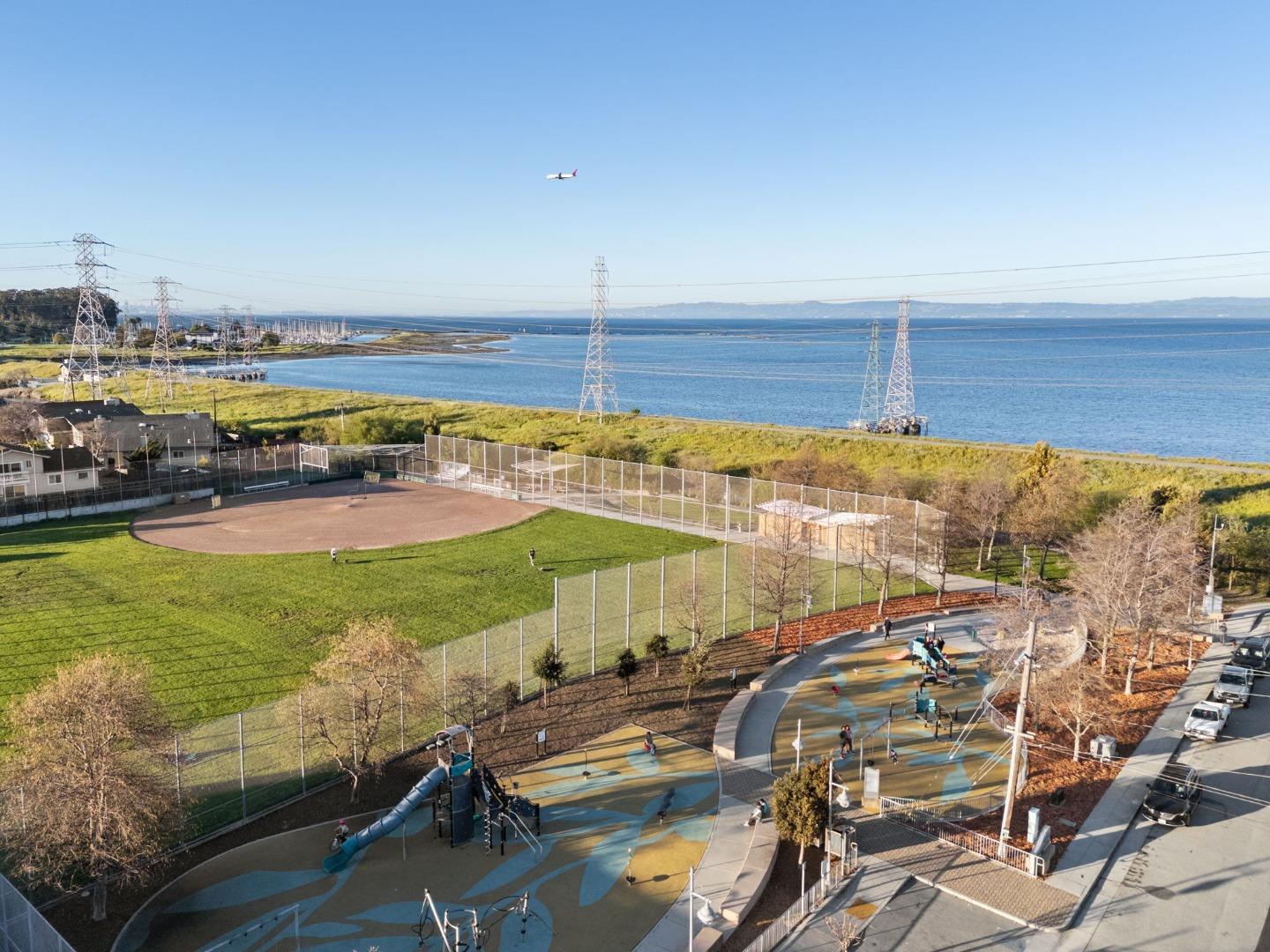 Bay view beyond a baseball field and playground, with power lines and a small airplane in the sky.