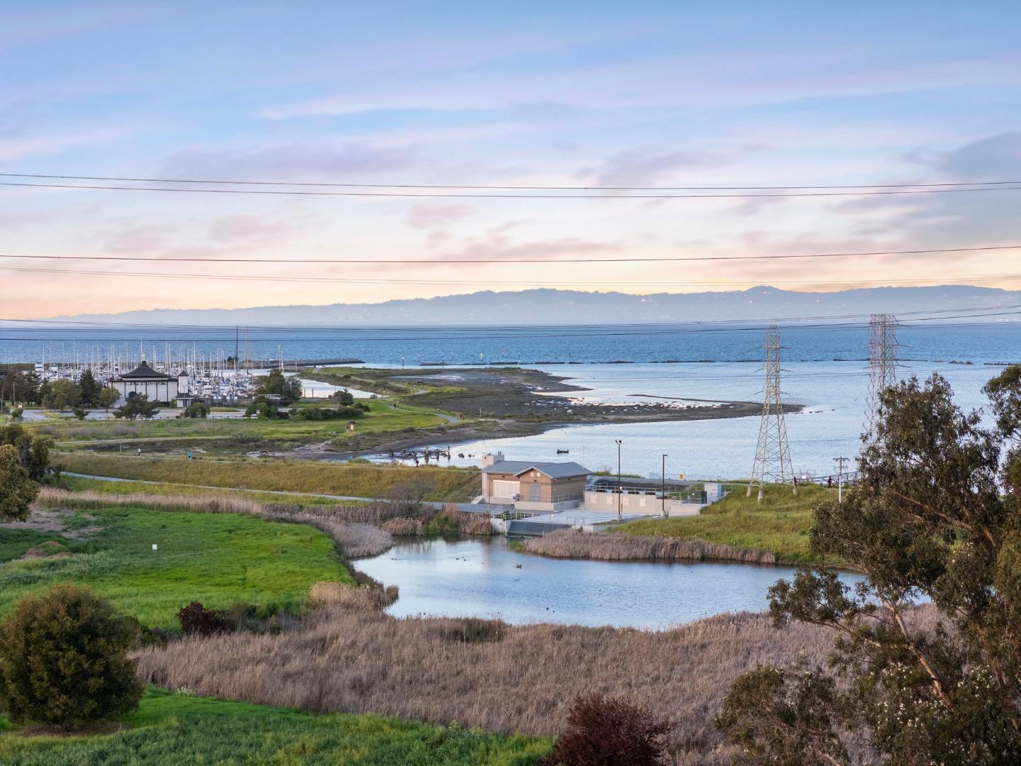 Waterfront view across marshland and a tidal inlet toward the bay, with a marina and distant hills.