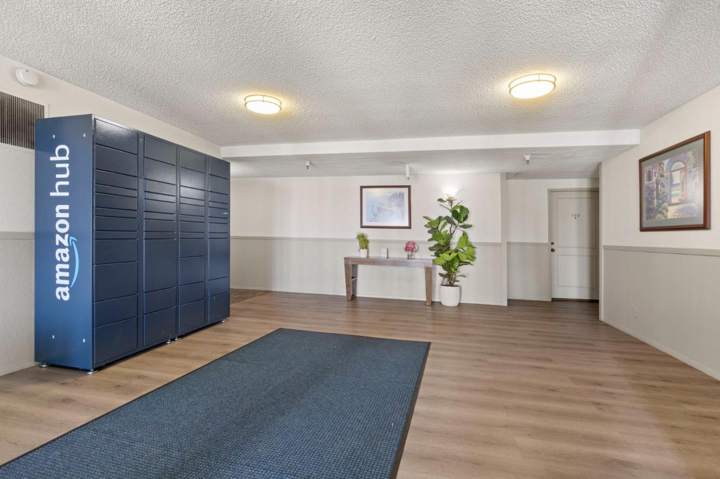 Apartment building lobby with Amazon Hub lockers, a blue runner, and a console table with plants.