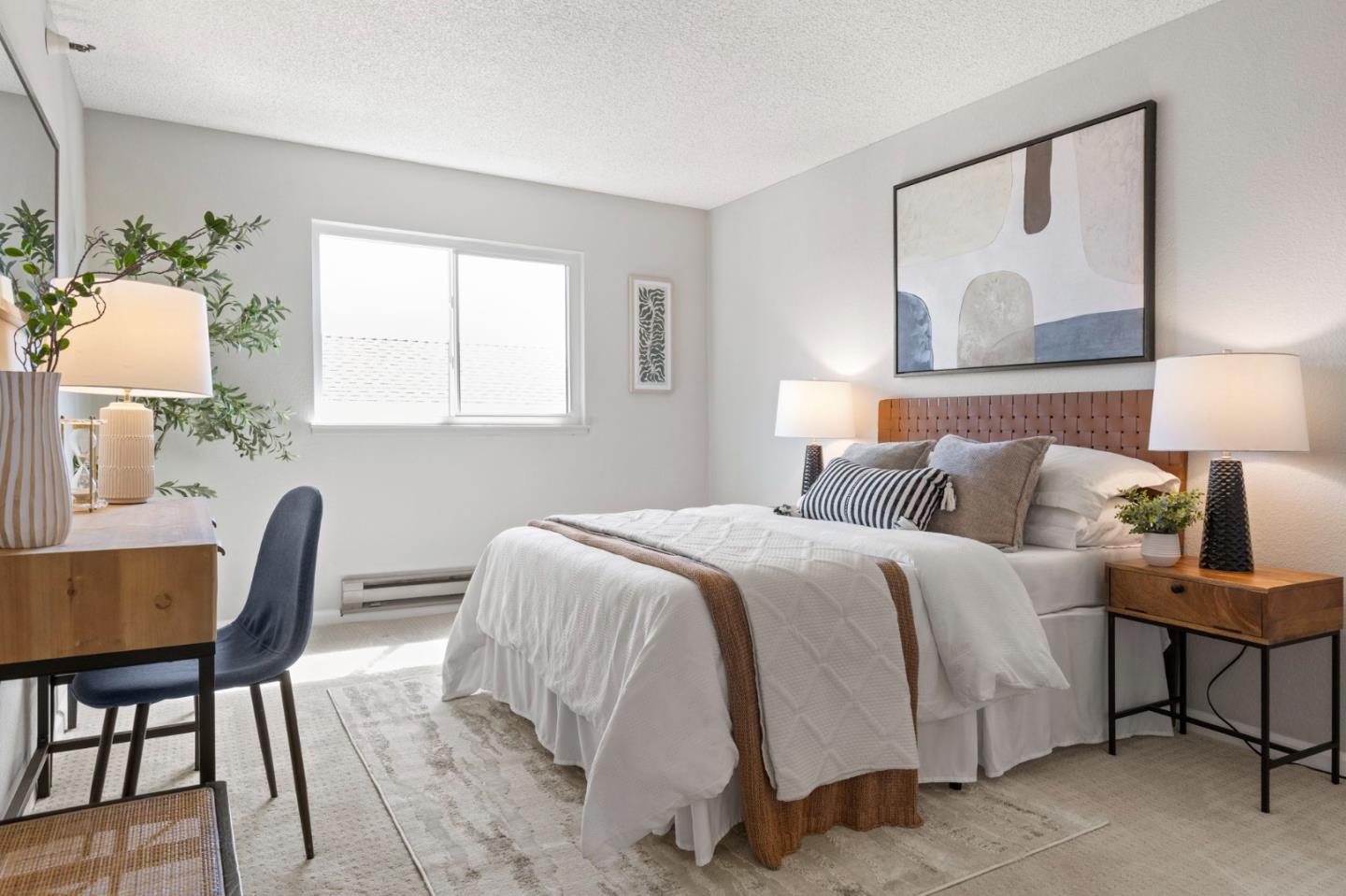 Staged bedroom with a white bed, wood headboard, abstract art, and a desk beside the window.