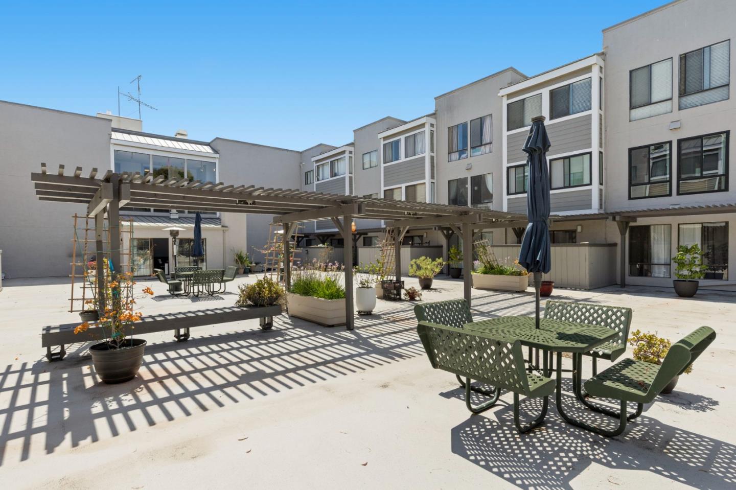 Residential courtyard with pergolas, green outdoor seating, and planters beside a multi-story building.