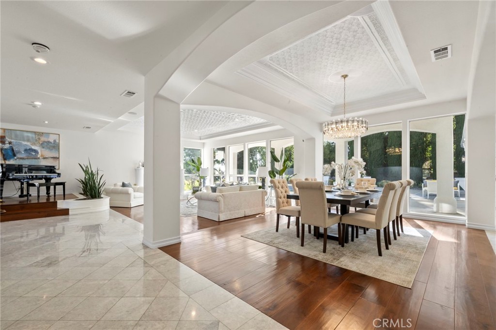 Open dining and living room with a tray ceiling, chandelier, and tall windows facing a green outdoor area.