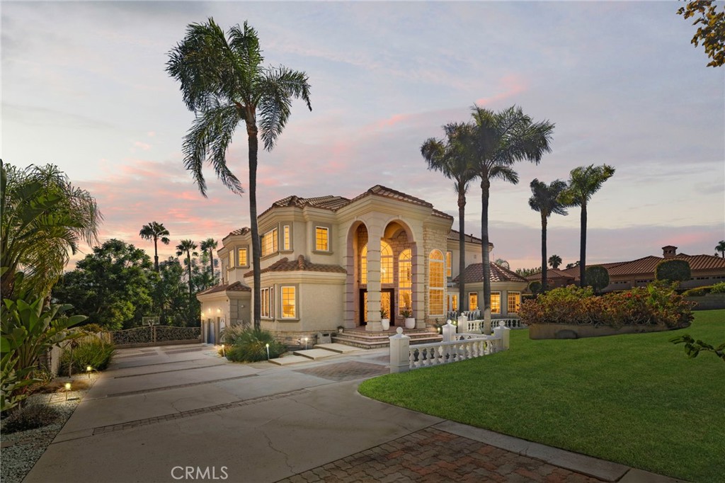 Mediterranean-style home with a tall arched entry, palm trees, and illuminated windows at dusk.
