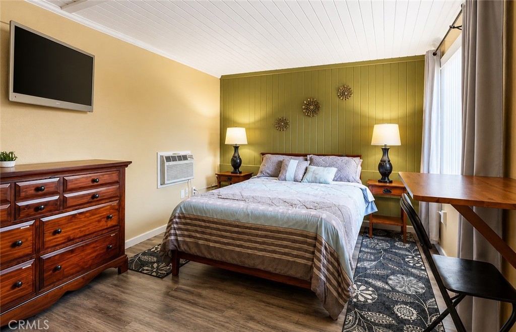 Bedroom with an olive-green paneled accent wall, large bed, wooden dresser, and desk beside a curtained window.