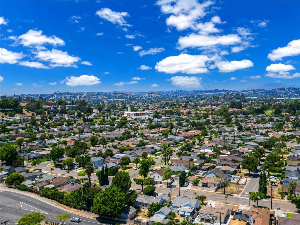 Aerial view of Monterey Park homes and tree-lined streets, with distant hills beneath a cloud-dotted blue sky.