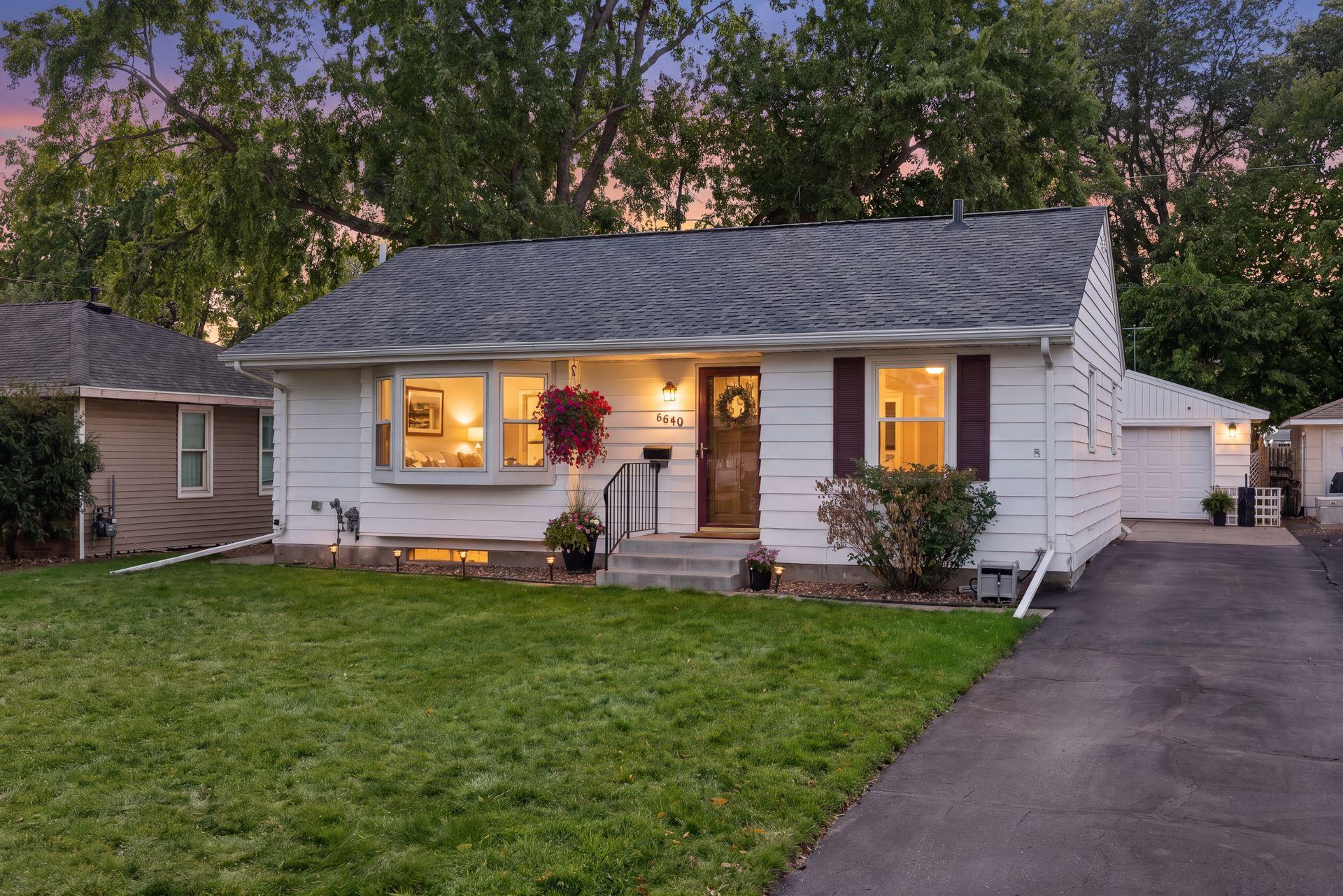 White single-story house with illuminated windows, flower planters, a front lawn, and driveway at dusk.