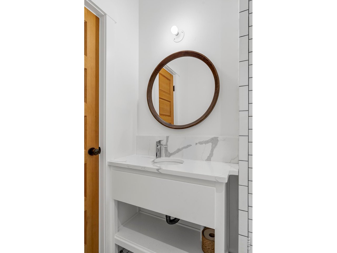 Bathroom vanity with a round wood-framed mirror, marble-look counter, and open shelving.
