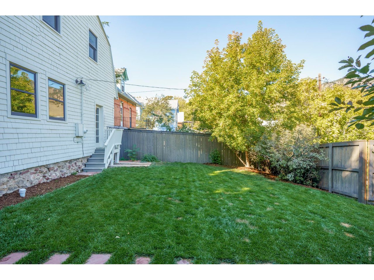 Backyard lawn beside a shingle-sided home, with steps, a privacy fence, and mature trees.