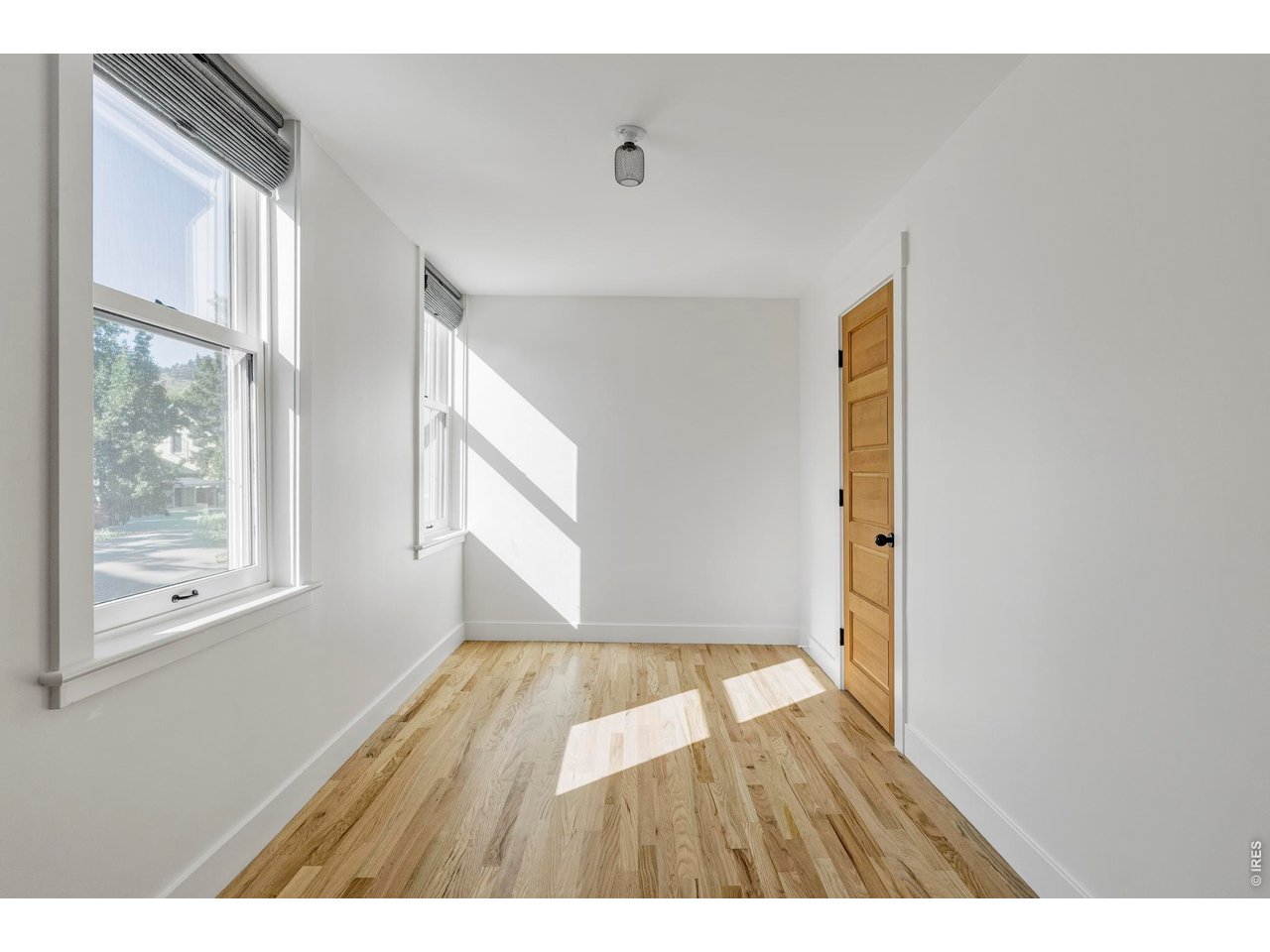 Bright empty room with large windows, light wood flooring, and a paneled wooden door.
