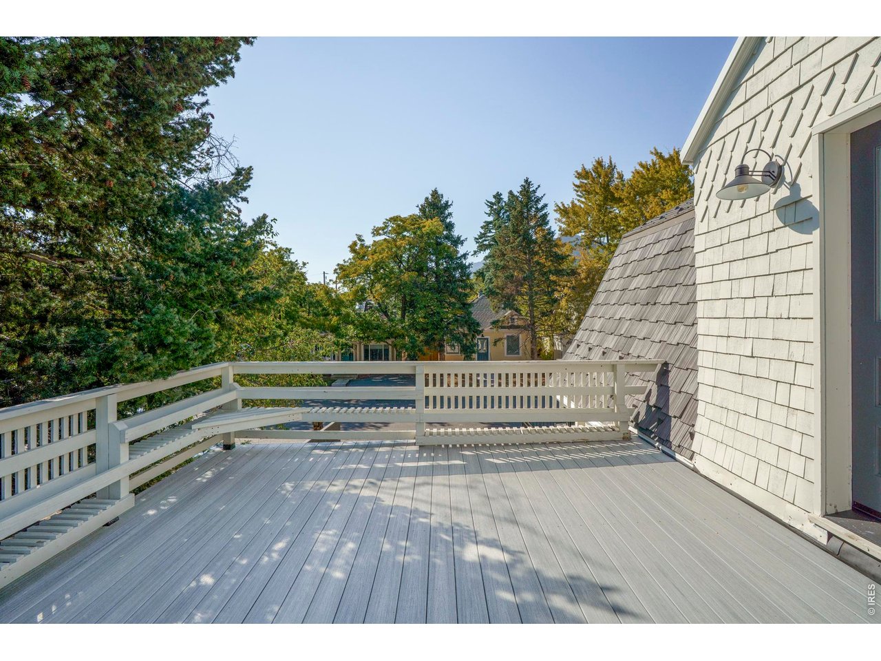 Spacious gray deck with white railings beside shingle siding, surrounded by leafy trees.