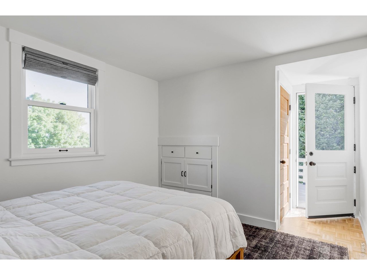 Bright bedroom with a white quilted bed, built-in cabinet, and open door to a sunlit exterior.