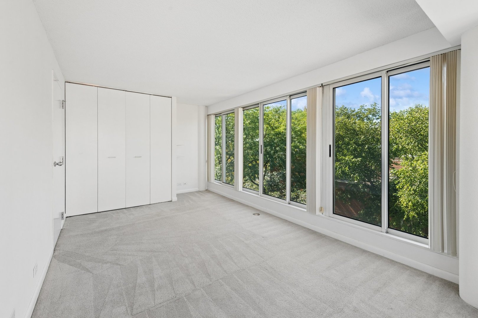 Empty bedroom with built-in closets and floor-to-ceiling windows overlooking trees.