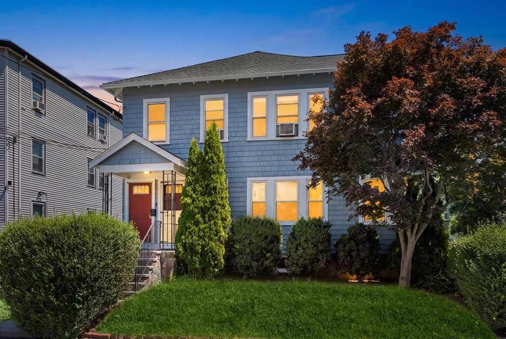 Blue two-story house with a red front door, glowing windows, and autumn tree at dusk.