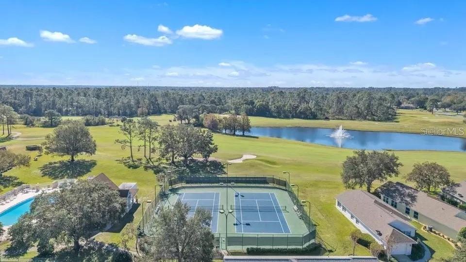Aerial view of a golf course, pond with fountain, tennis courts, and nearby homes.