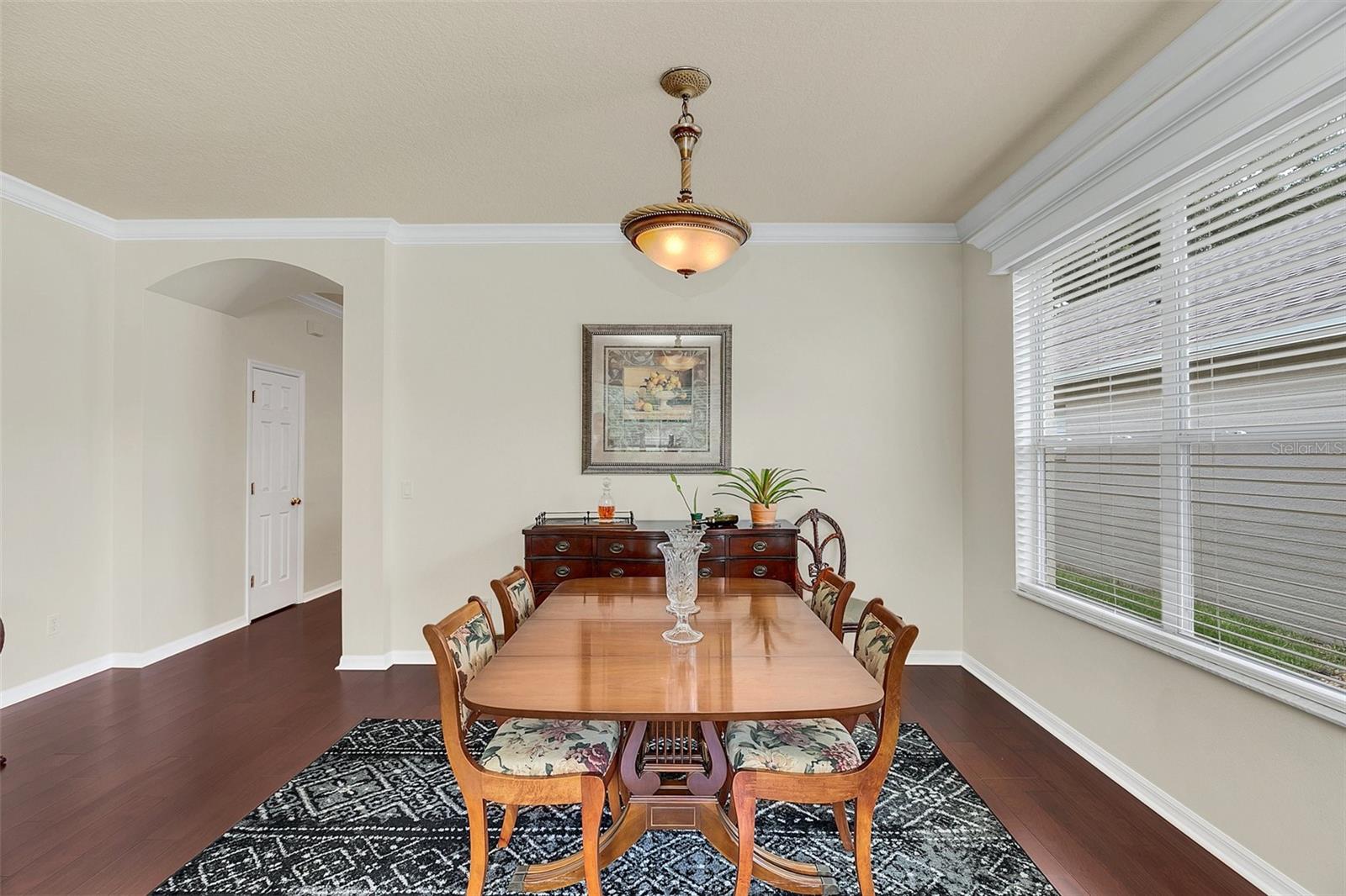 Dining room with a long wooden table, floral-upholstered chairs, and a large window with blinds.