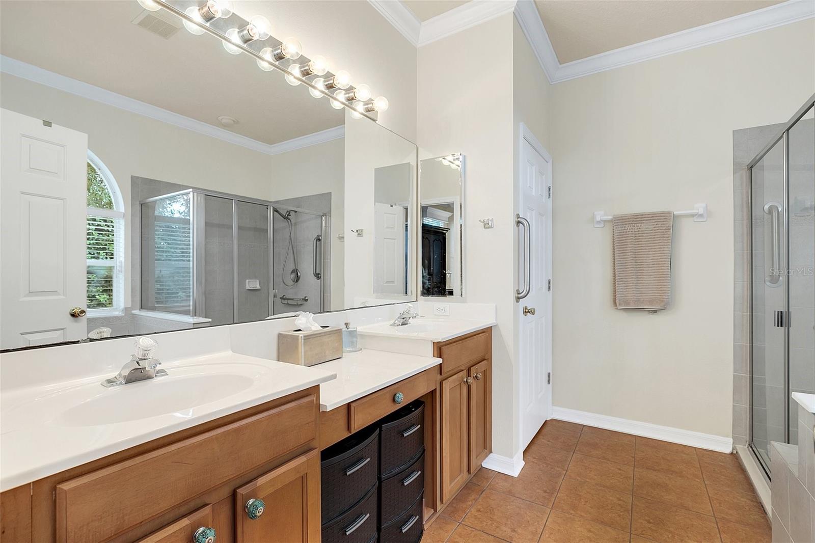 Bathroom with a wood double vanity, expansive mirrors, arched shuttered window, and glass-enclosed showers.