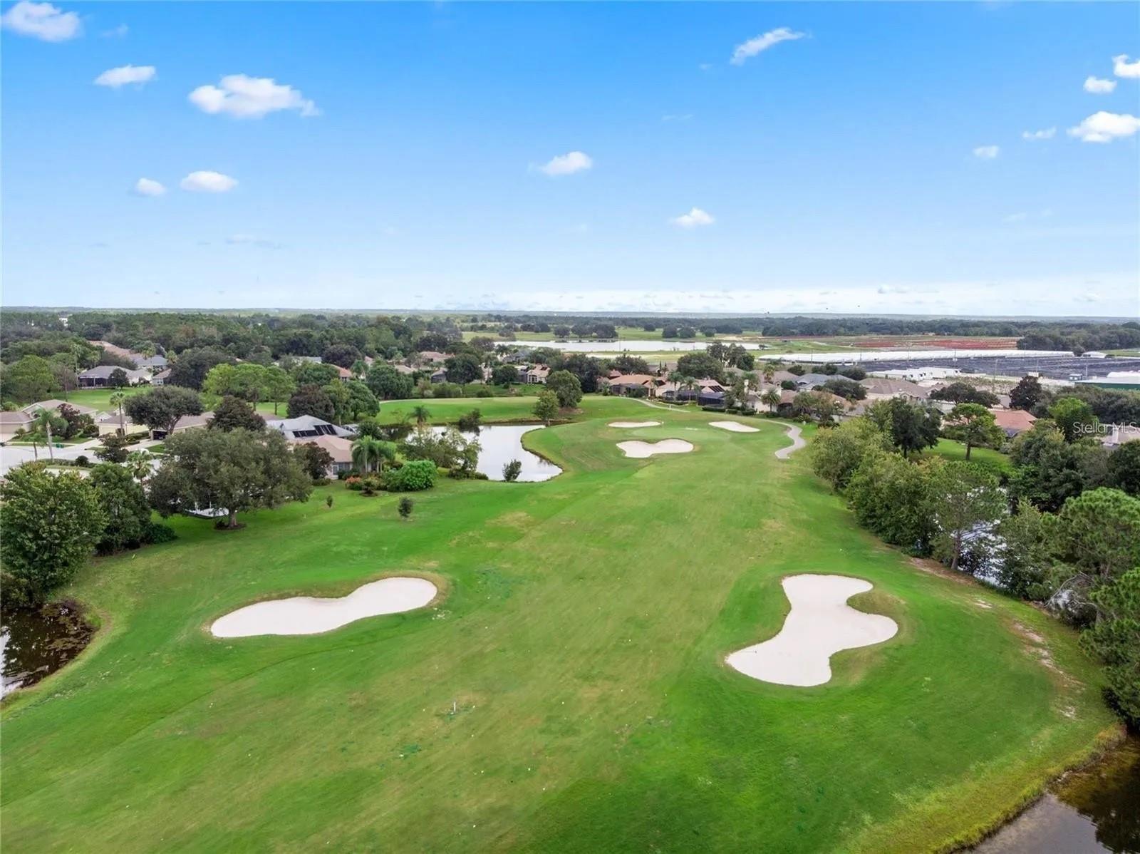 Aerial view of a golf course with sand bunkers, a pond, and homes beyond.