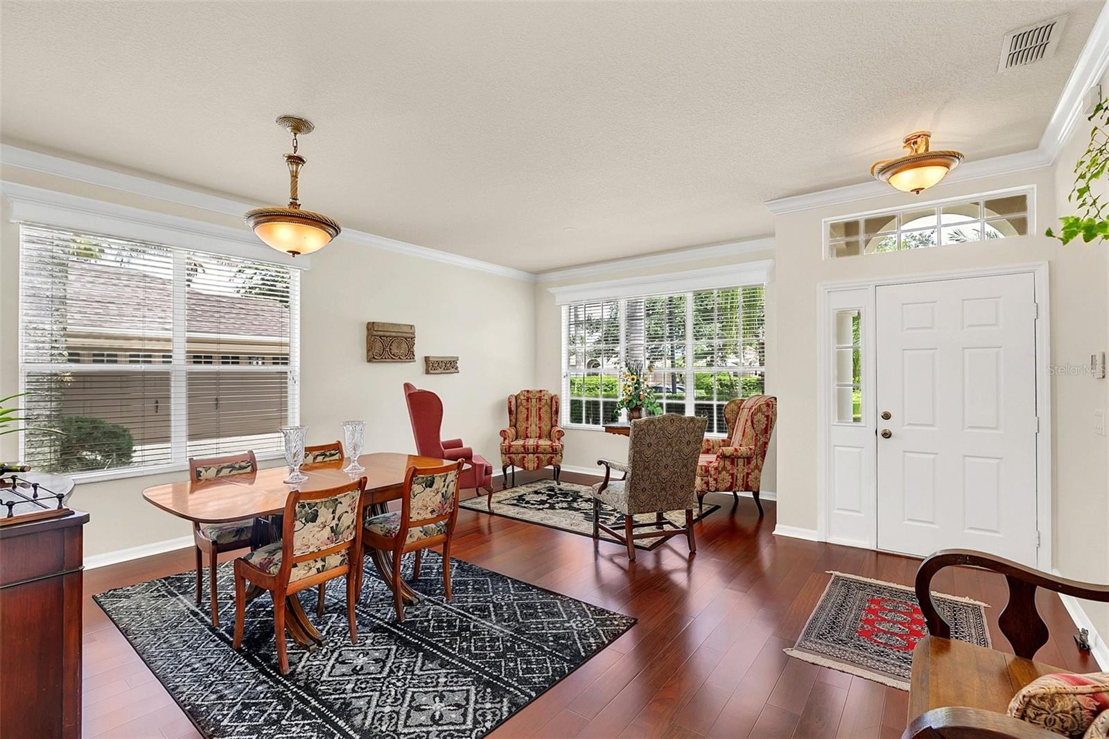 Furnished dining and sitting area with polished wood floors, large windows, and a white double front door.