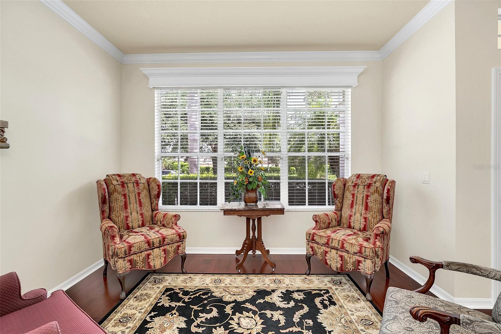 Sitting area with patterned armchairs, a flower-topped table, and a large window with white blinds.