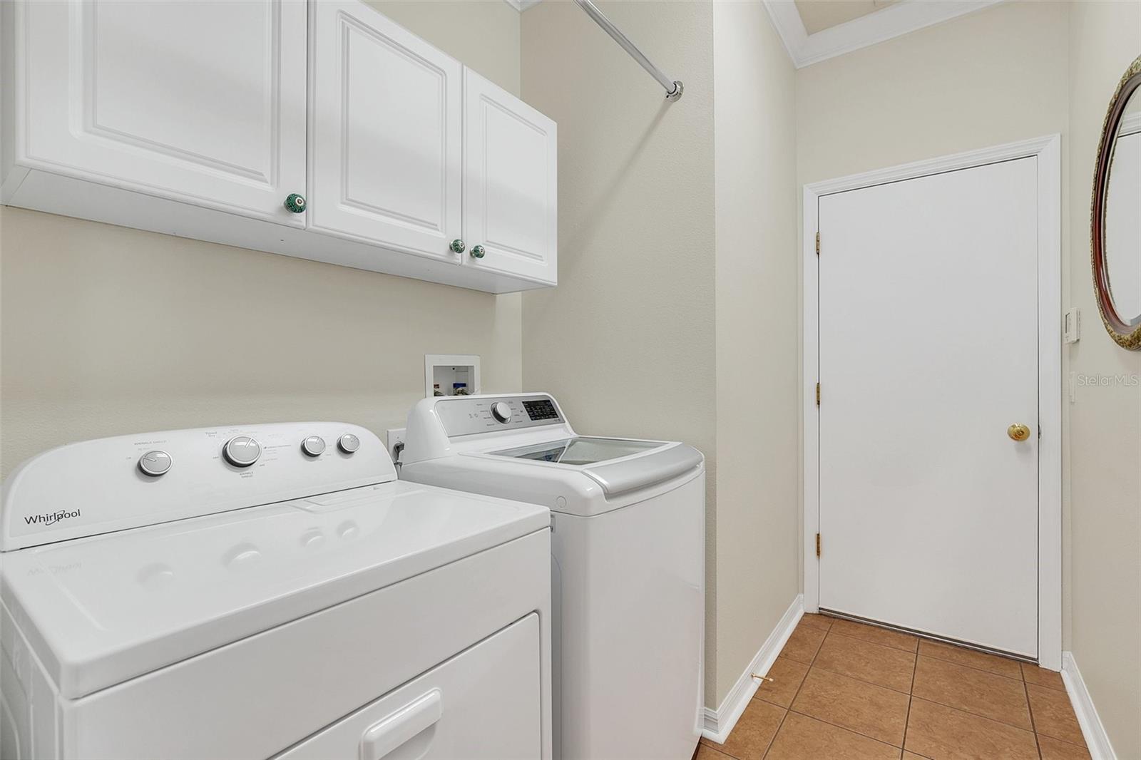 Laundry room with white washer and dryer beneath upper cabinets, with tan tile flooring.