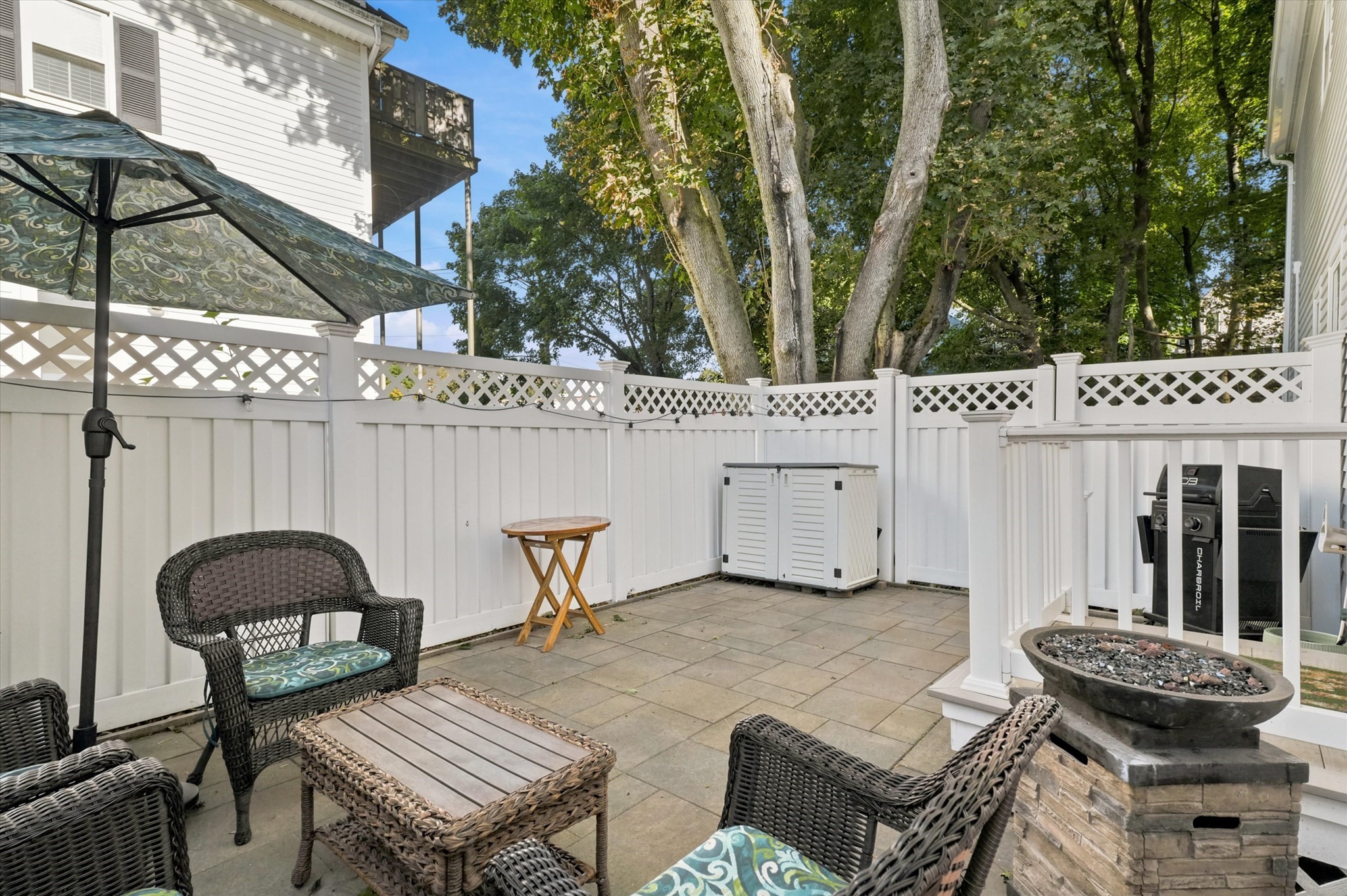 Furnished patio with wicker seating, a patterned umbrella, white fencing, and mature trees.