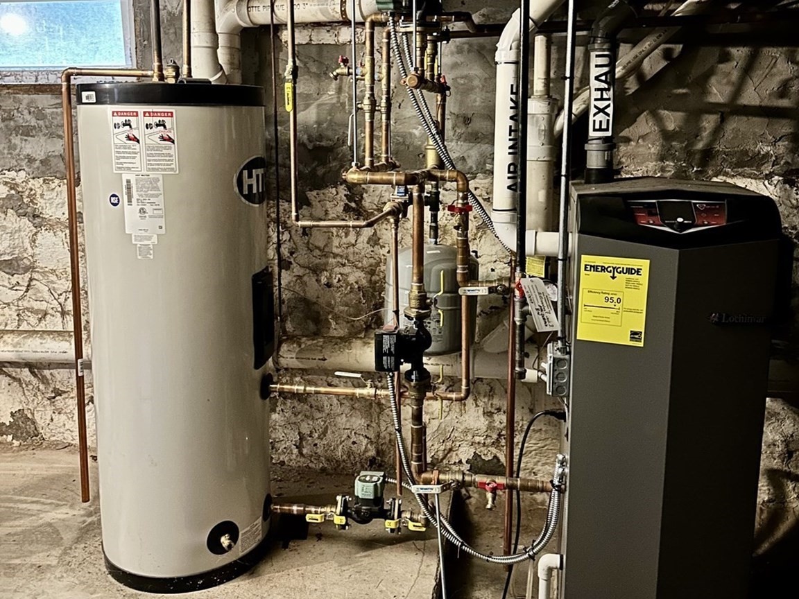 Basement mechanical room with a water heater, boiler, and exposed piping against a stone foundation.