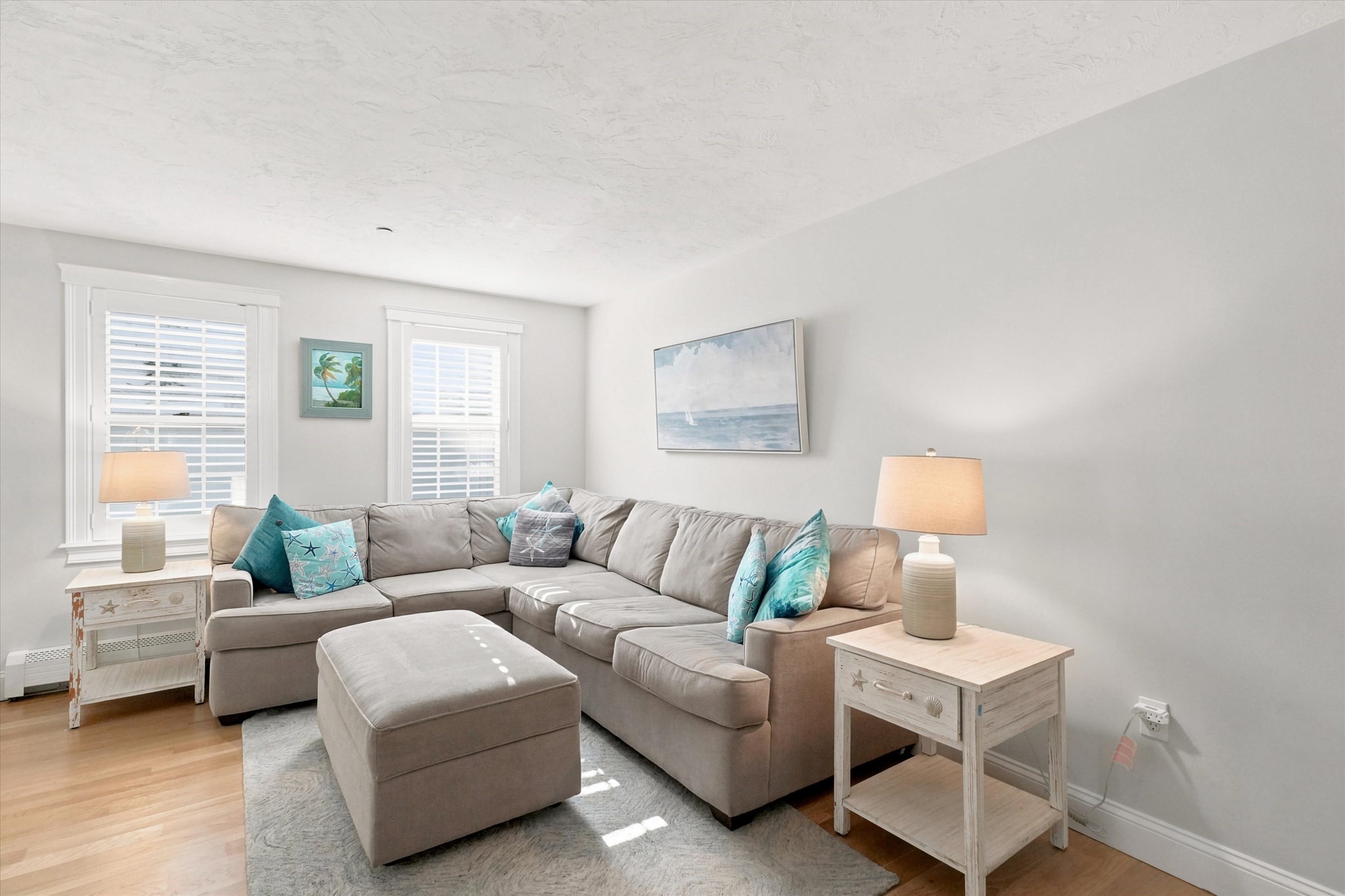 Living room with a gray sectional, turquoise pillows, shuttered windows, and coastal artwork.