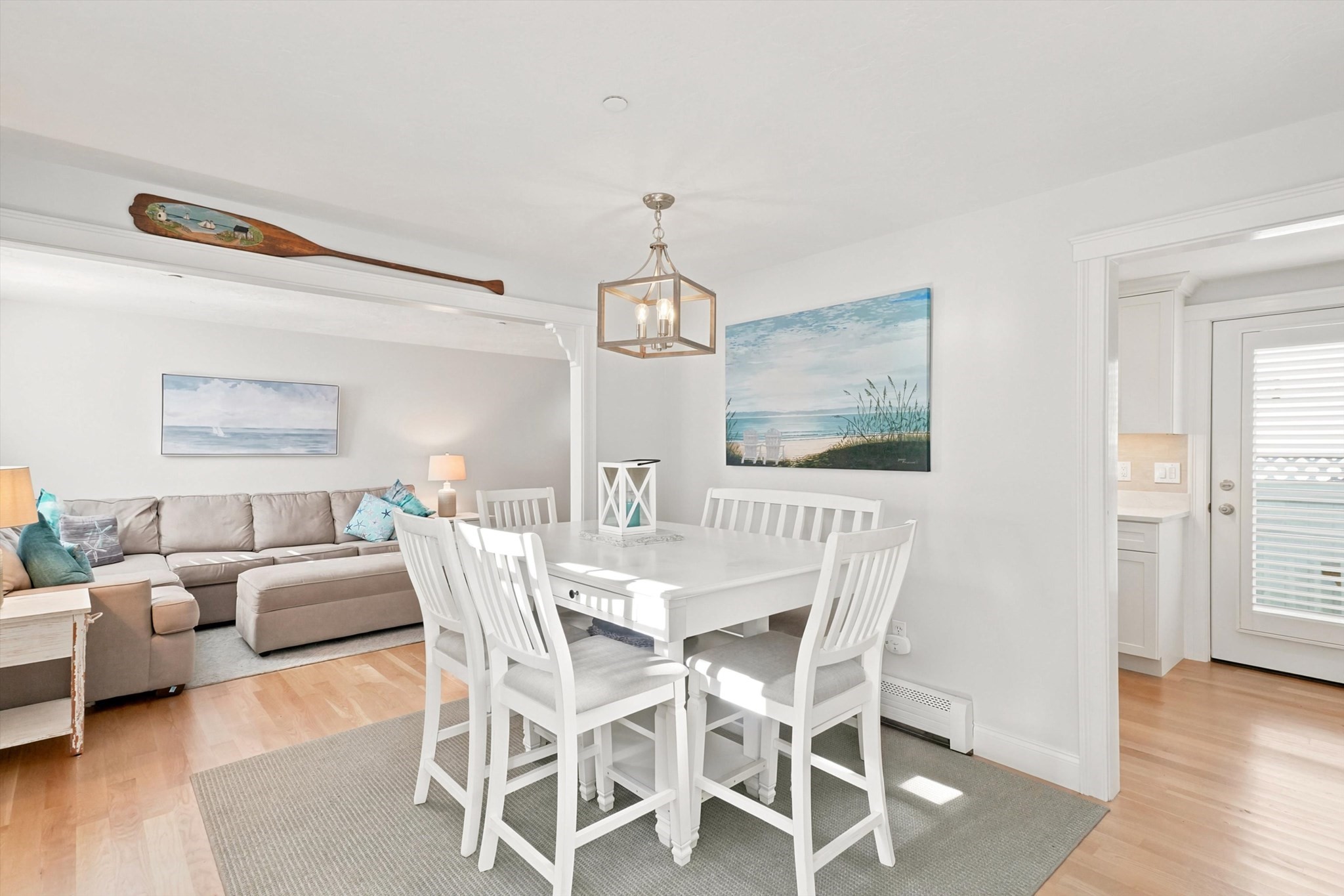 Open-plan dining and living area with white furnishings, a beige sectional, and coastal artwork.