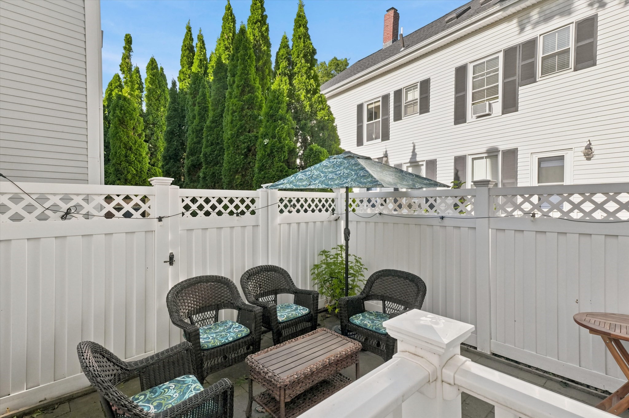 Fenced patio with wicker seating, patterned cushions, and an umbrella beside a white-sided home.