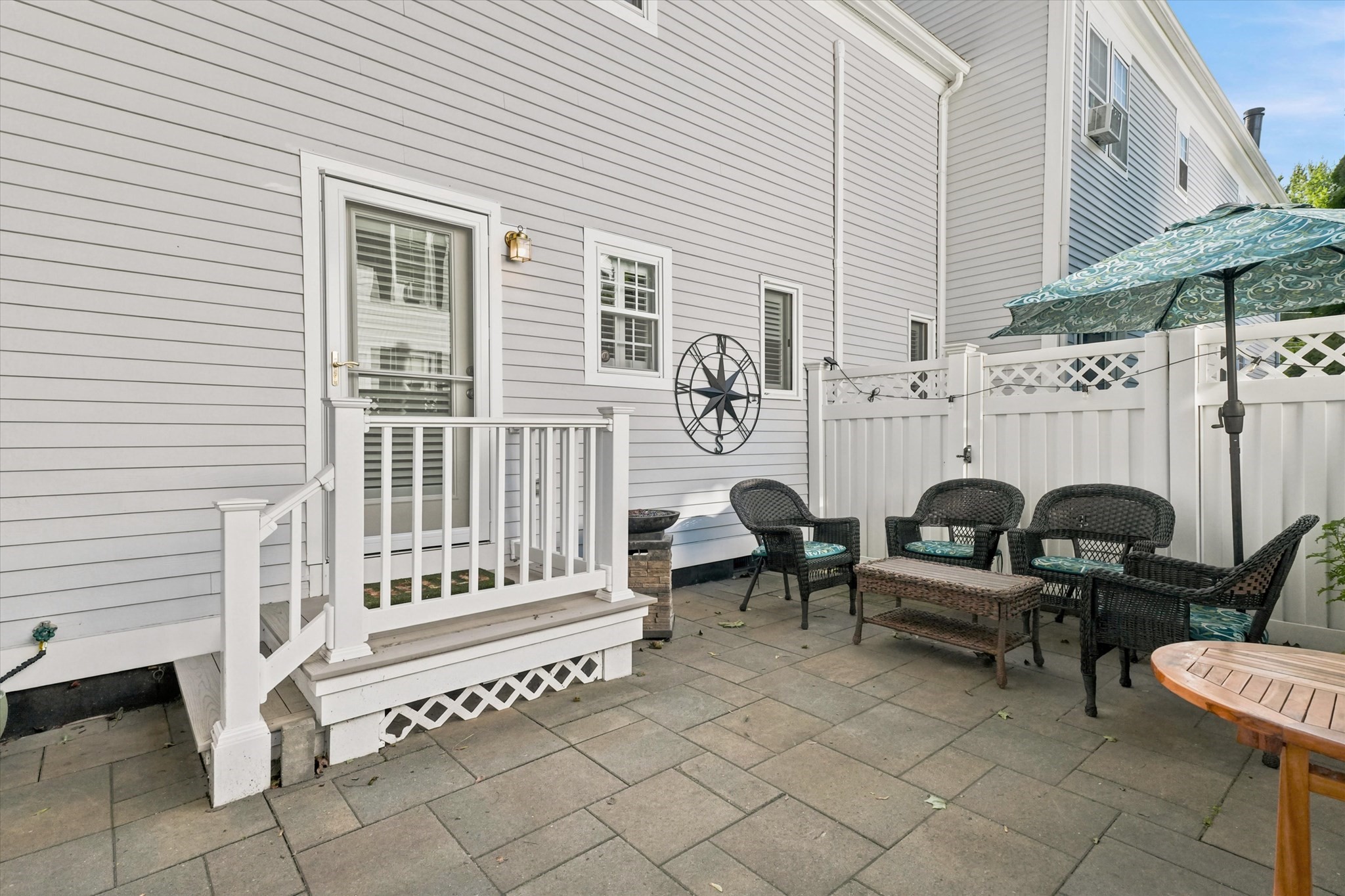 Paved patio with wicker seating, a patterned umbrella, and a small white-railed entry deck.
