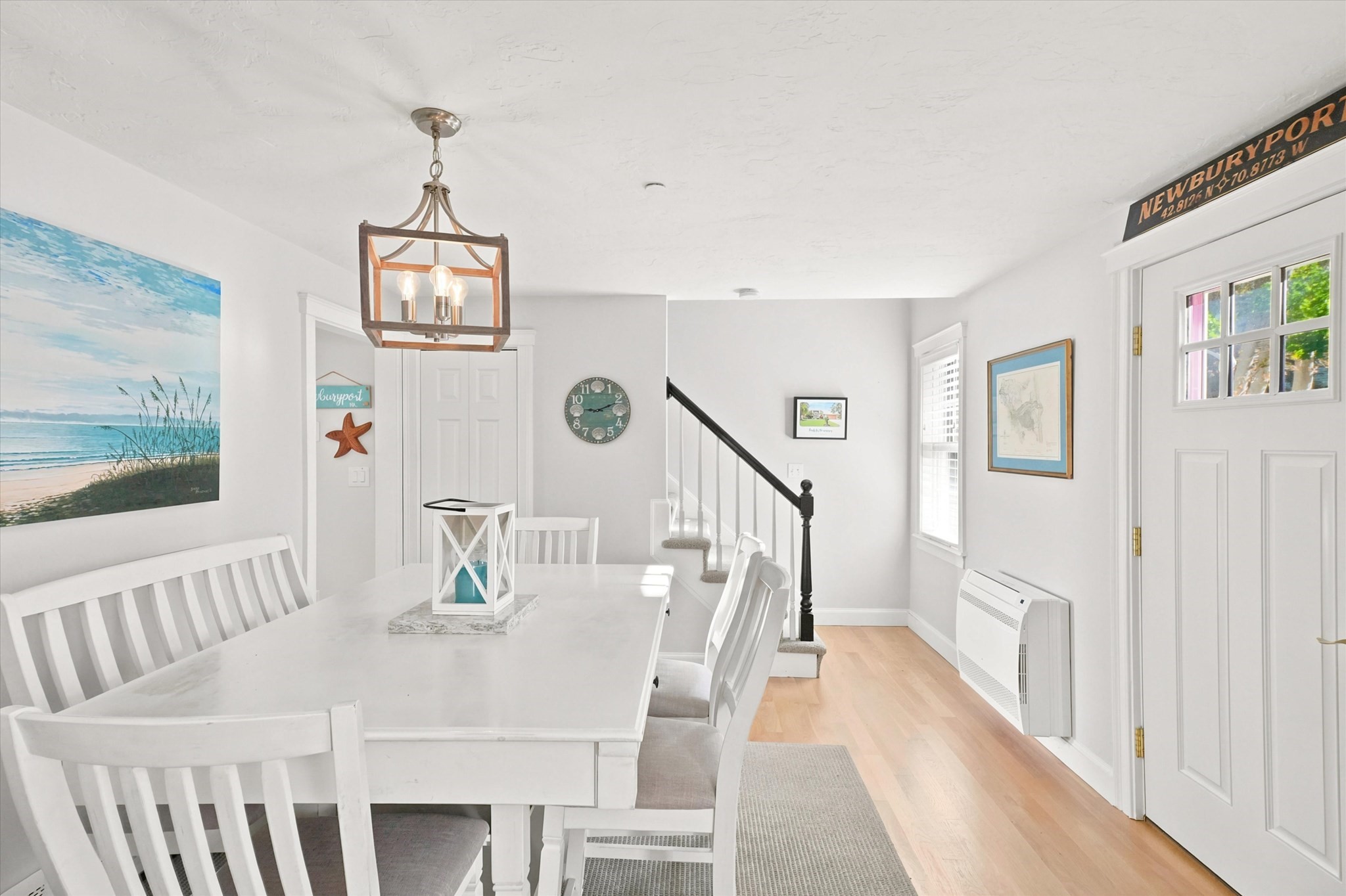 Dining area with a white table, coastal artwork, pendant light, and staircase with black railing.